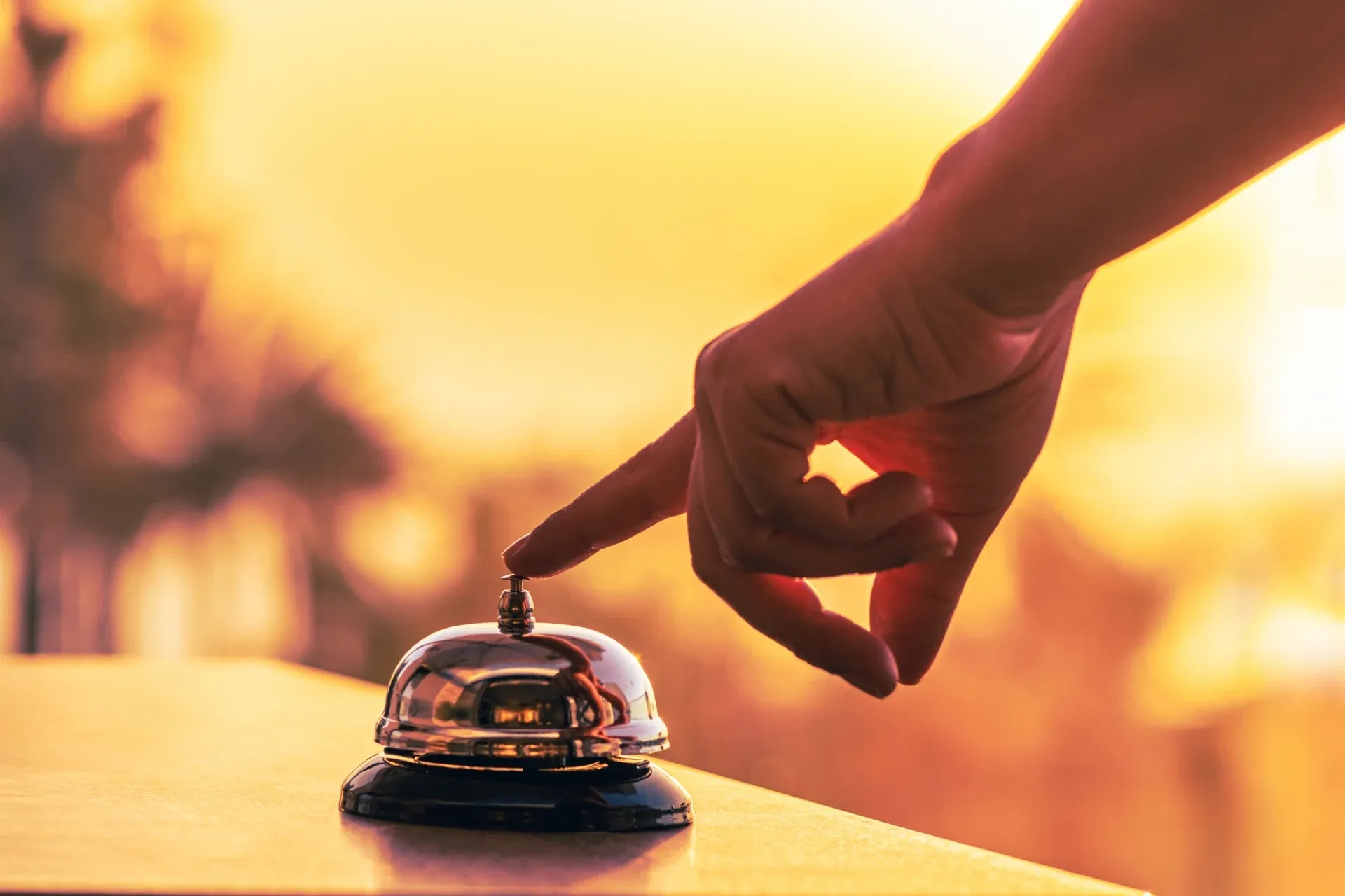 A hand poised to press a shiny service bell against a warm, blurred sunset background. The image conveys anticipation and a sense of calm service.