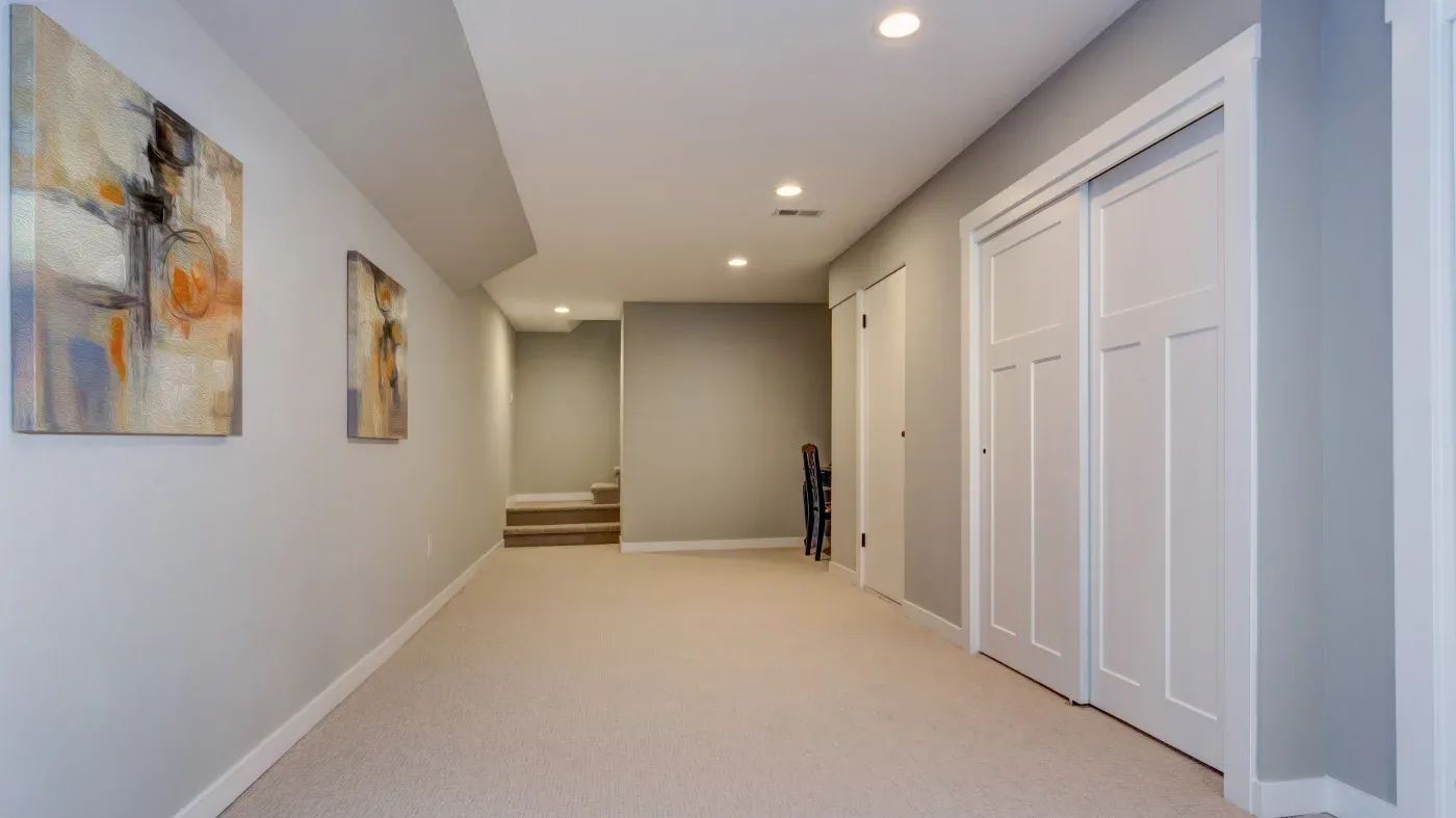 A bright, spacious hallway with beige carpet and gray walls. Abstract art hangs on the left, while white closet doors and a staircase are on the right.