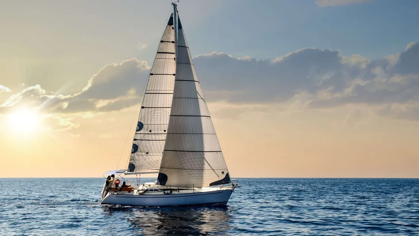 A sailboat glides on a calm, blue ocean under a clear sky with a setting sun. The scene conveys serenity and freedom.
