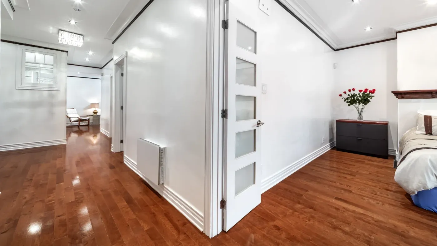 A modern interior with white walls and polished wooden floors. A corridor leads to a bright living area with a chair. A bedroom has a dresser with flowers.