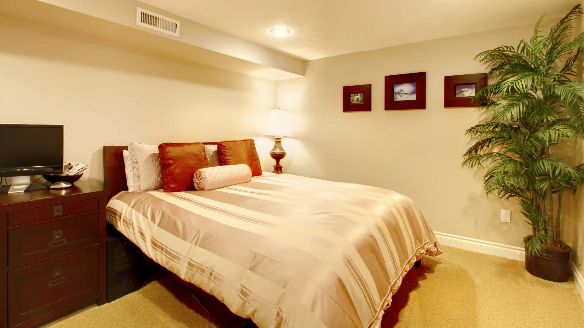 A cozy bedroom with a beige striped bedspread, pillows, and a rolled cushion. A lamp casts a warm light. A potted plant and framed photos adorn the space.