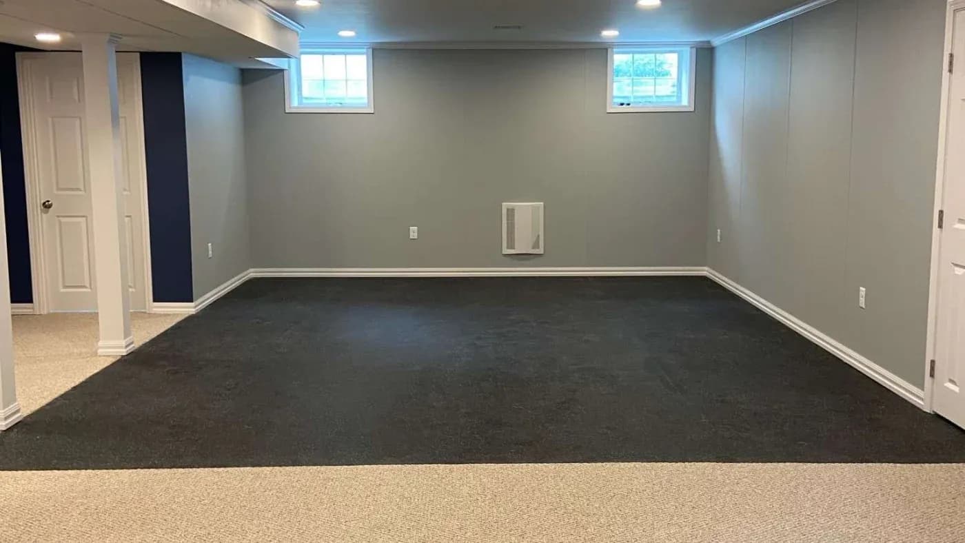 A spacious basement with light gray walls and contrasting dark carpet at the center. Natural light from two small windows creates a calm ambiance.