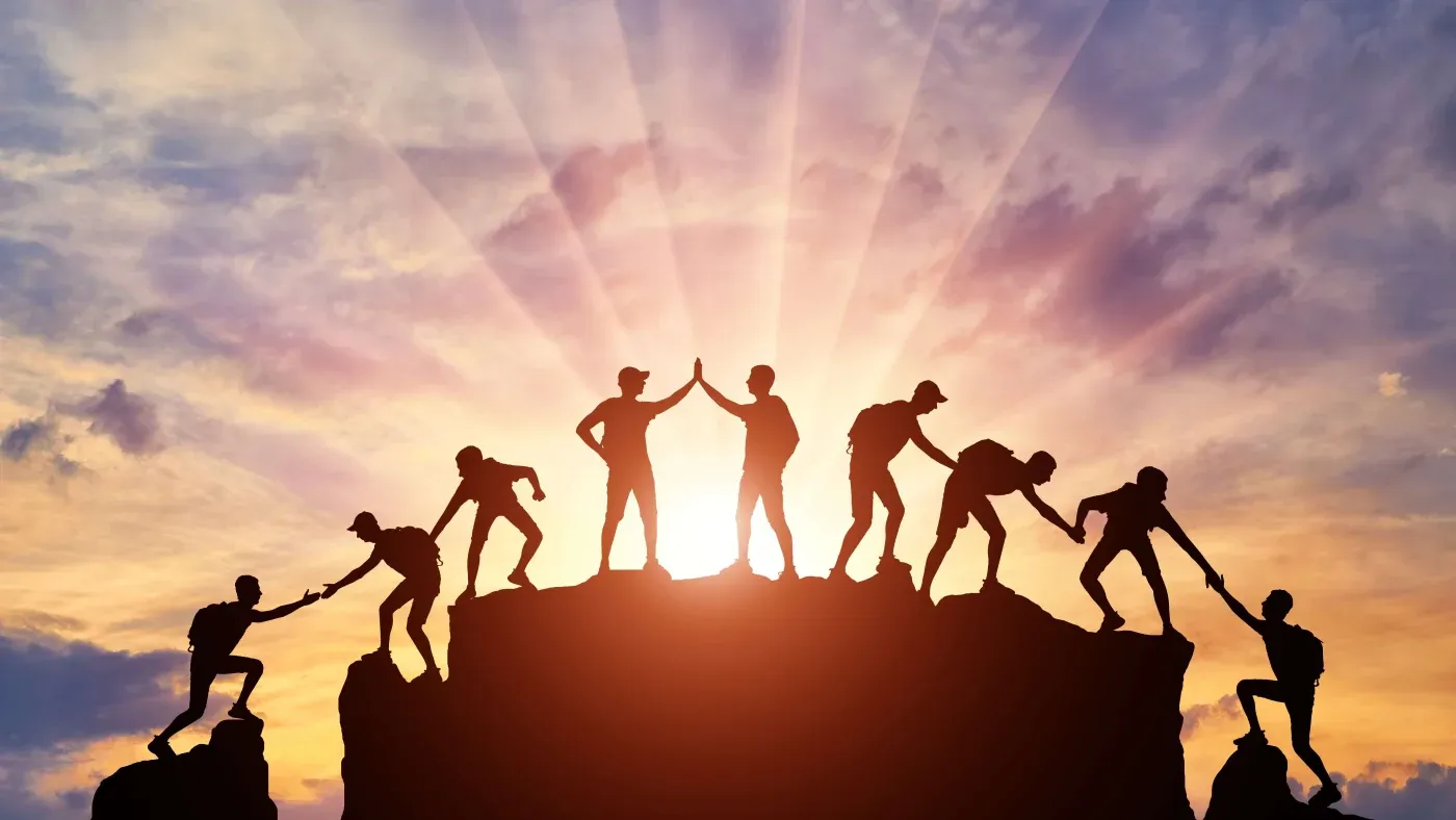 Silhouetted figures climb a mountain against a vibrant sunrise sky, helping each other. The scene conveys teamwork, support, and achievement.