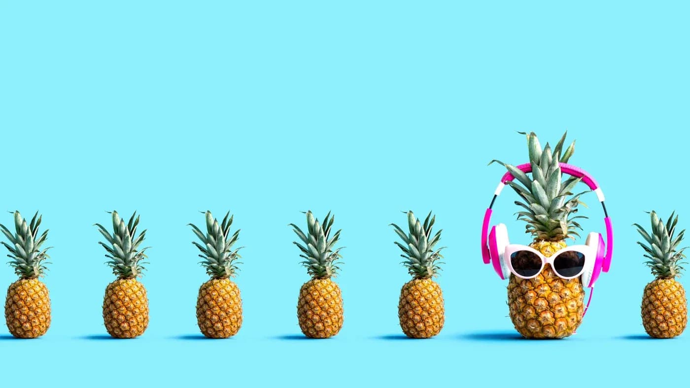A row of six pineapples on a blue background. The last pineapple wears sunglasses and pink headphones, adding a playful, quirky touch.