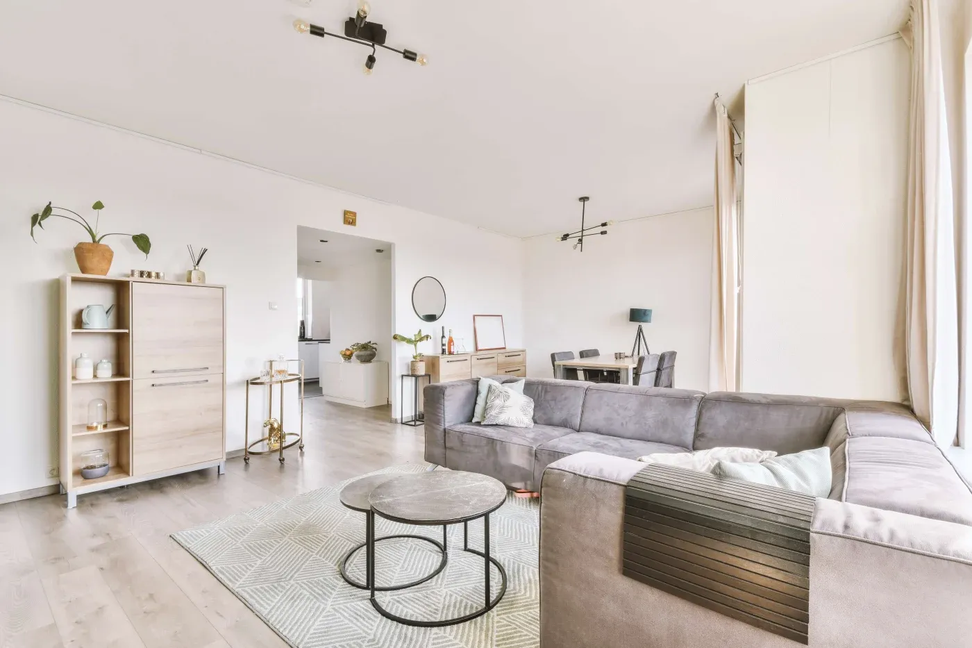 Modern living room with light wood flooring, featuring a large gray sectional sofa and round coffee tables on a patterned rug. Minimalist decor includes shelves with plants and ceramics, a dining area with a round mirror, and soft natural lighting from large windows. Neutral tones create a calm and inviting atmosphere.