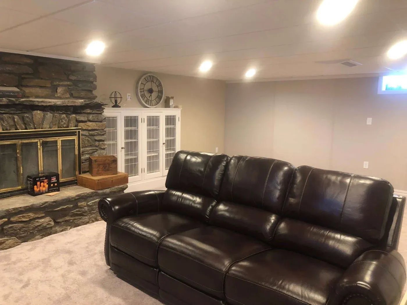 Cozy basement living room with a brown leather sofa, stone fireplace, and glass-front cabinets. Soft lighting creates a warm, inviting atmosphere.