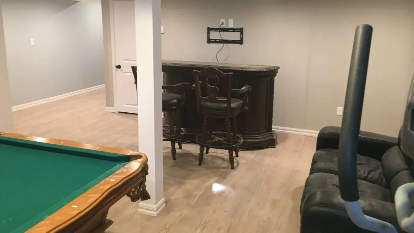A basement room features a pool table with a green top, a wooden bar with two stools, a black leather couch, and an exercise machine on a wooden floor.