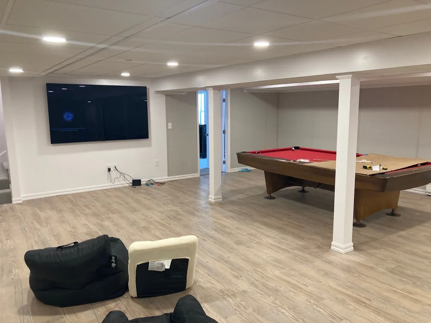 Modern basement with light wood flooring, large wall-mounted TV, red-felt pool table, upright chair, and cozy bean bags. Bright, relaxed atmosphere.