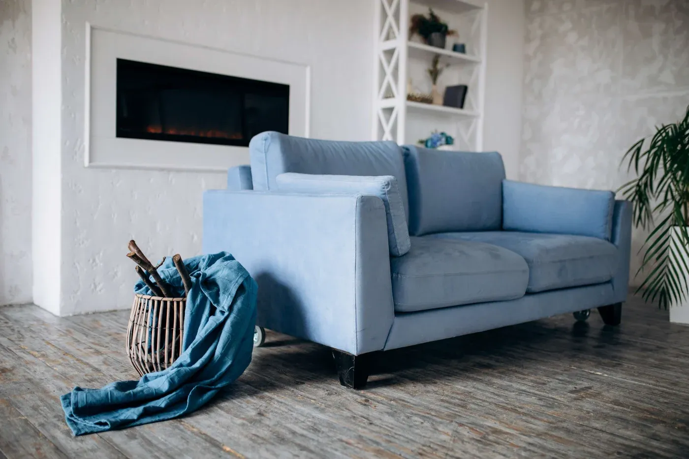 A cozy living room features a soft blue sofa on a wooden floor, a wicker basket with a draped fabric, and a modern black fireplace in the background.