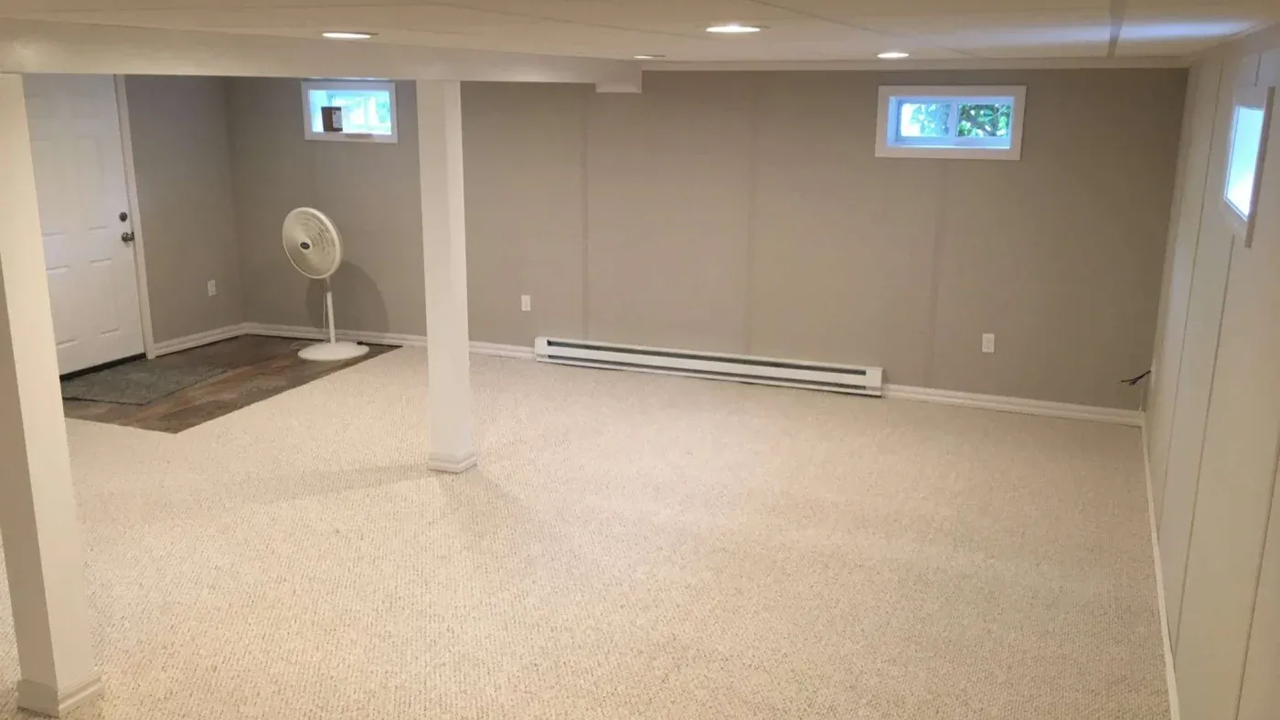 A spacious, empty basement with light beige carpeting and neutral walls. A white oscillating fan stands in the corner near a door. Recessed lighting adds a warm ambiance.