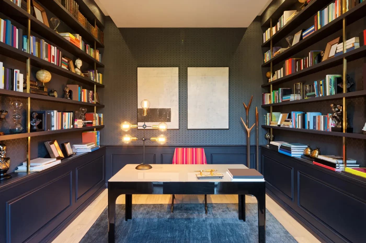 Modern home office with dark walls, bookshelves filled with books and globes on either side. Central sleek desk with a unique lamp, striped chair, and two abstract paintings on the wall, creating a sophisticated and focused atmosphere.