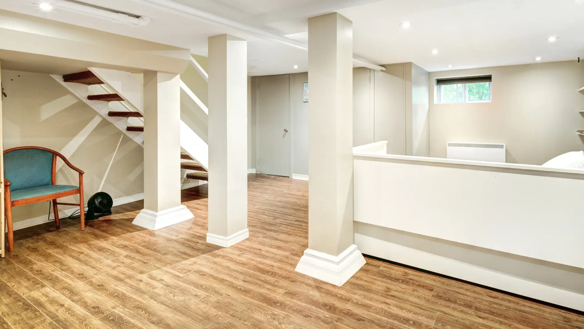 Spacious basement with light wood flooring, white walls, and ceiling columns. A wooden chair sits under the stairway, creating a clean, minimalistic feel.