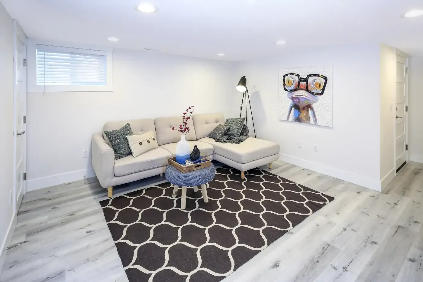 Modern living room with a beige sectional sofa and assorted cushions, a stylish floor lamp, a geometric rug, and whimsical frog artwork. Bright and inviting.