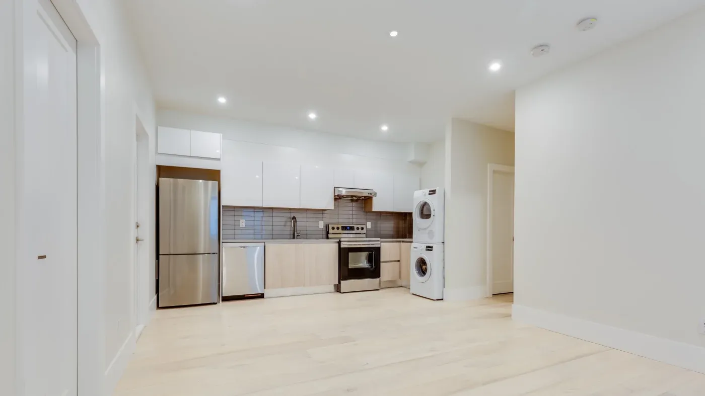 Modern kitchen with white cabinets, stainless steel appliances, and a stacked washer-dryer. Bright, minimalist space with light wood floors.