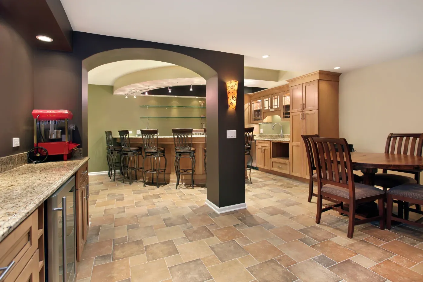 Spacious basement bar with stone tile flooring, wooden cabinetry, and a cozy dining area. A popcorn machine sits on a granite counter, creating a warm, inviting ambiance.