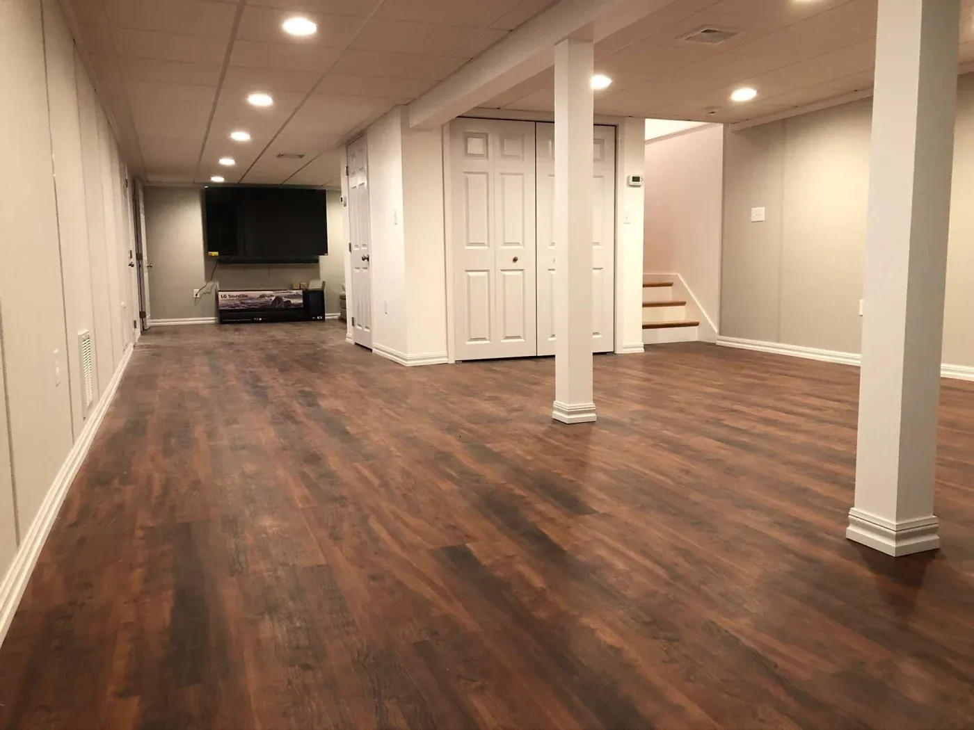 Spacious basement with polished wood flooring, white walls, and ceiling lights. A TV is mounted in the background, and a staircase is on the right.