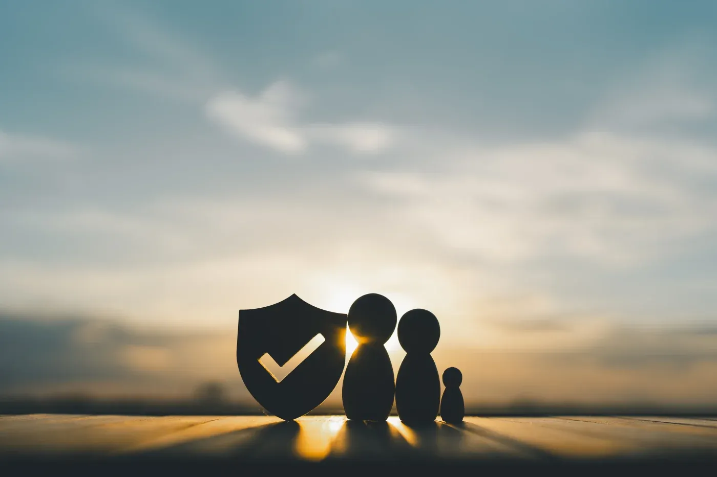 Silhouettes of three wooden figures and a shield with a checkmark on a table, set against a peaceful sunrise sky, symbolizing family protection.