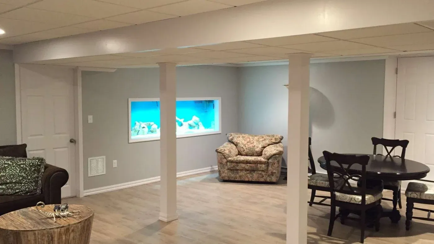 Spacious basement with a large wall aquarium, floral armchair, round dining table with chairs, and wood flooring, creates a cozy and inviting atmosphere.
