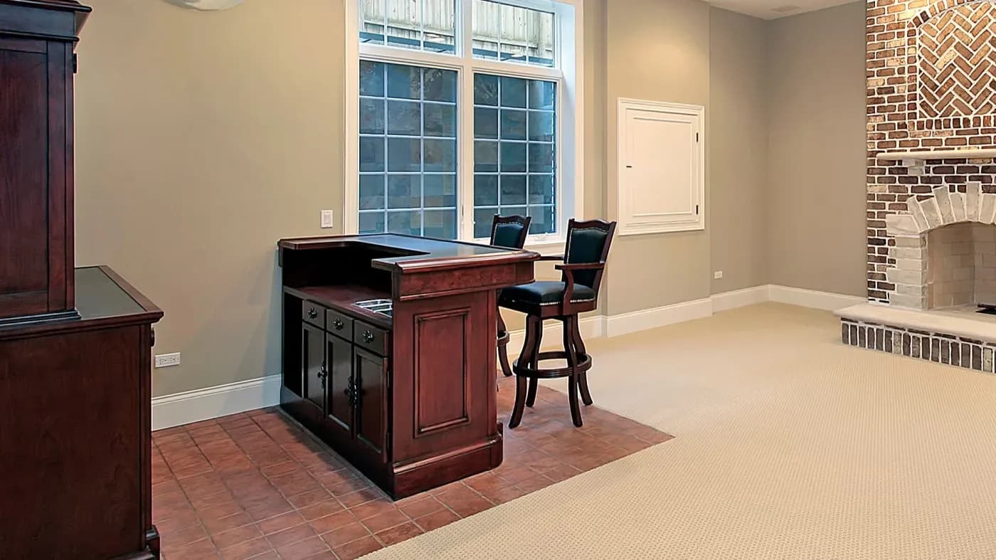 Warm room with a wooden bar and two high chairs on a tiled floor. Large window, beige carpet, and a brick fireplace create a cozy, inviting atmosphere.