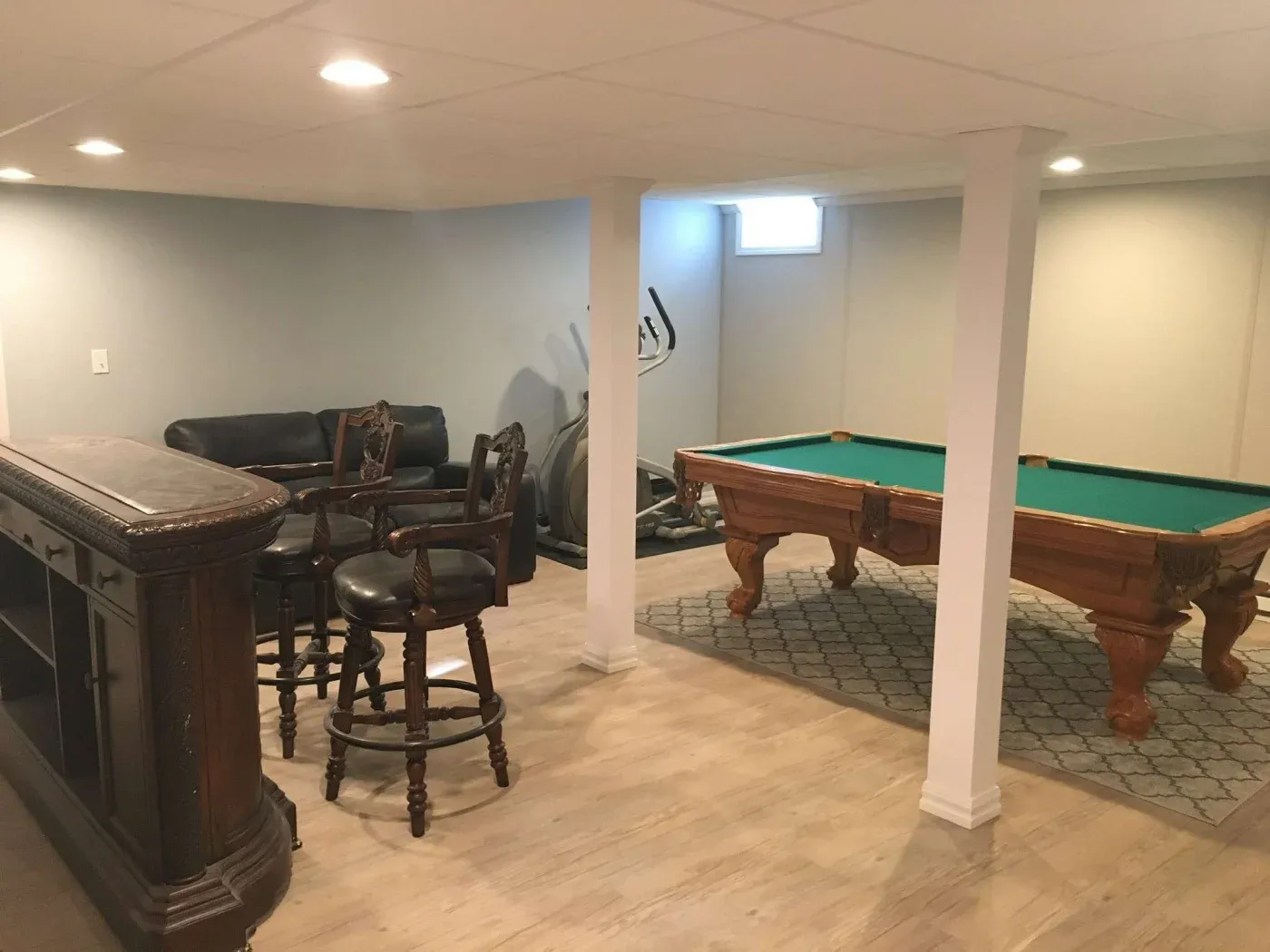 A basement game room with pool table on a carpet, wooden bar with stools, black leather sofa, and exercise equipment. Warm, inviting atmosphere.