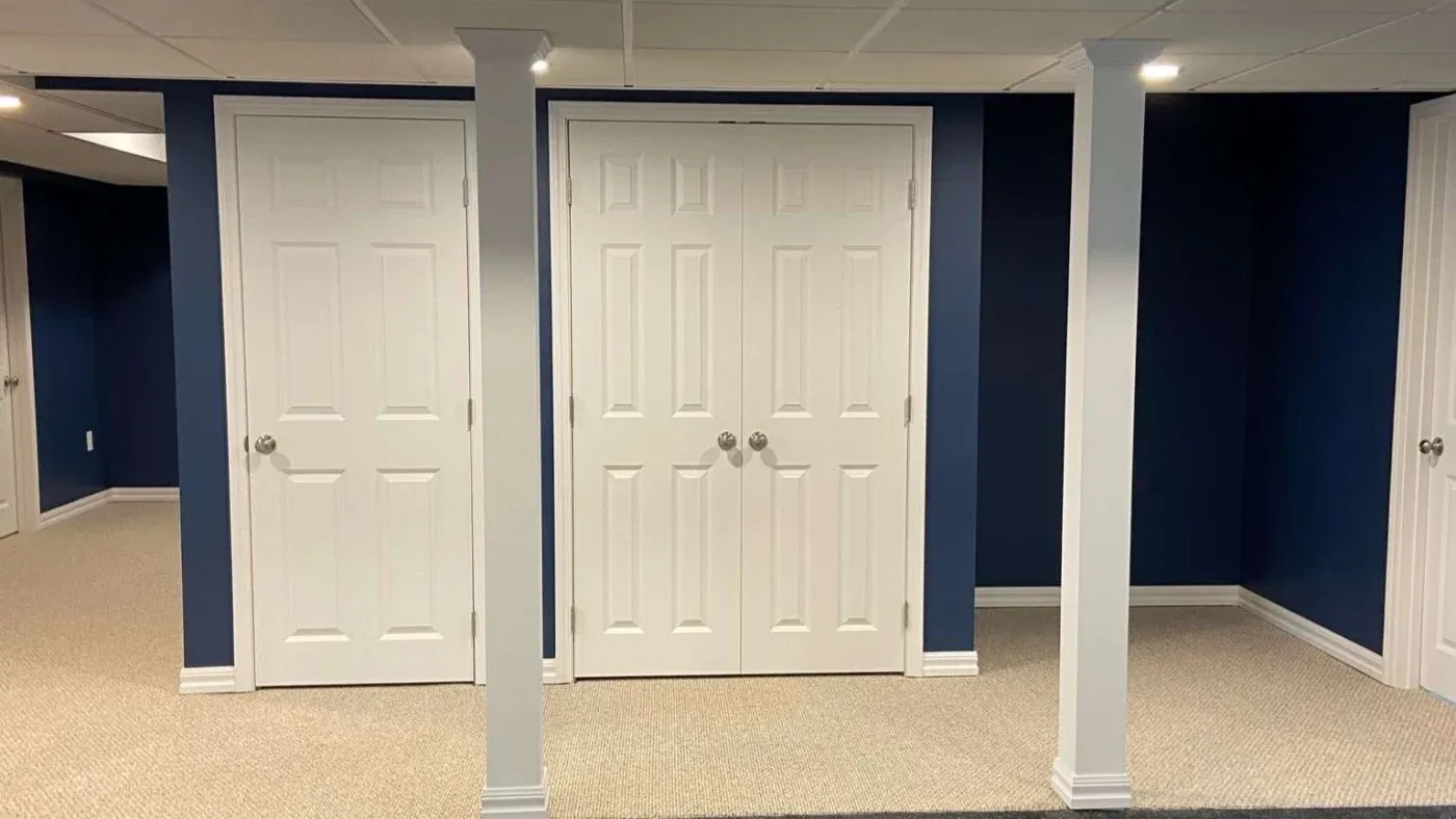 Basement room with dark blue walls, beige carpet, white ceiling, and three white doors. Two supporting columns stand in the foreground, adding symmetry.
