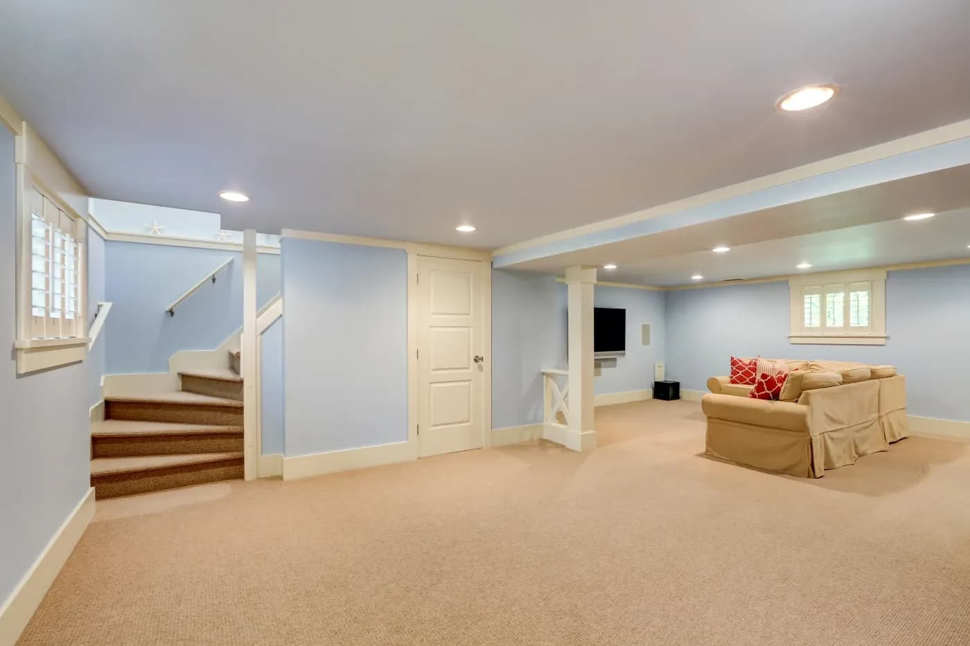 Spacious, carpeted basement with light blue walls, beige sectional sofa, red pillows, and a wall-mounted TV. Stairs lead up, creating a cozy, inviting vibe.