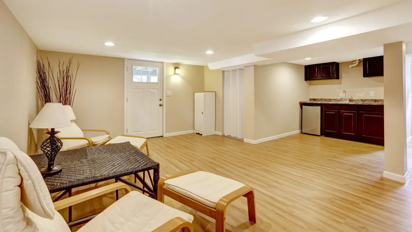 Spacious basement with light wood flooring, beige walls, and recessed lighting. Features cozy seating, a wicker table, and a kitchenette with dark cabinets.