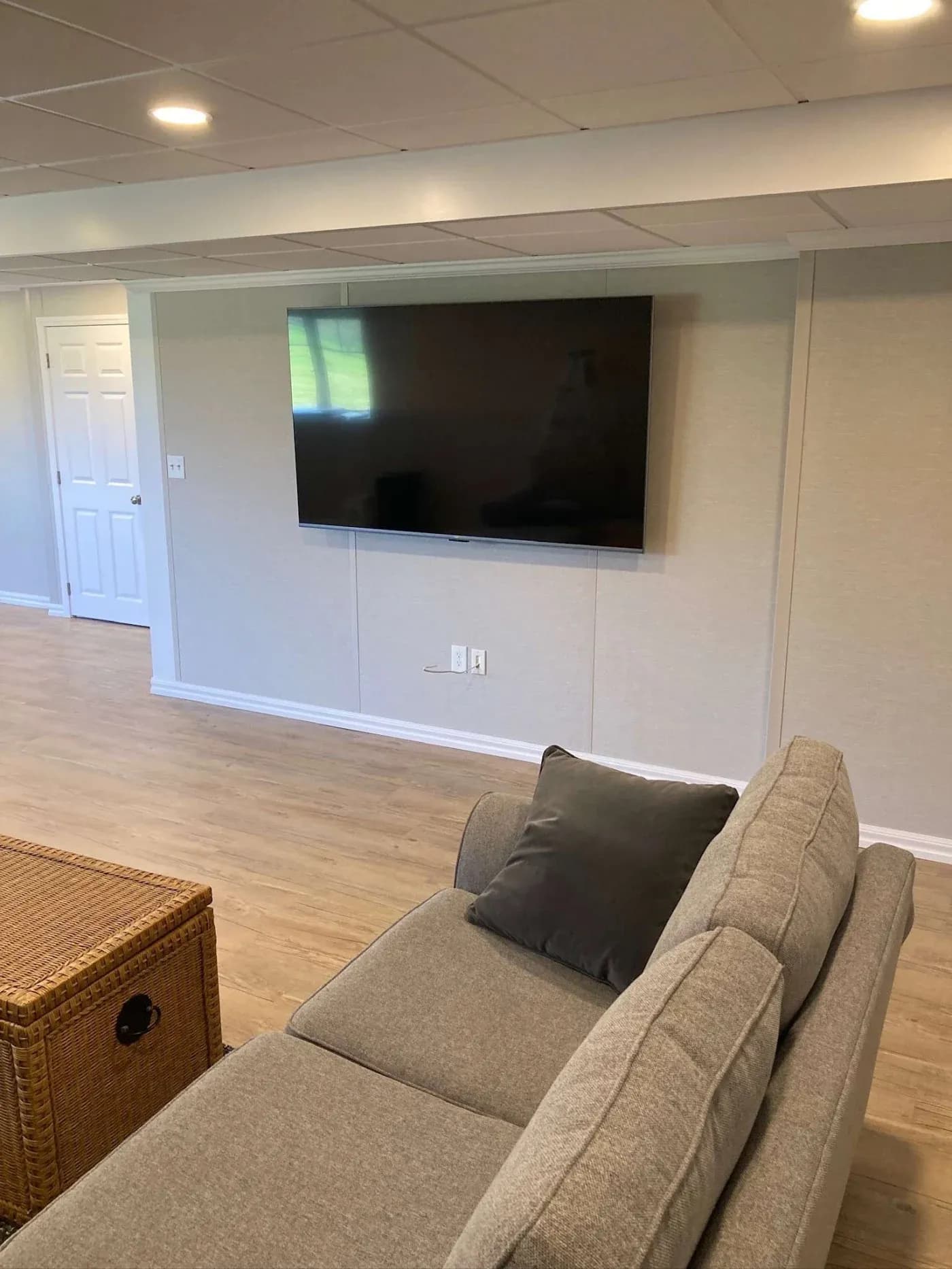 A modern living room with a large flat-screen TV mounted on a beige wall. A gray sofa with dark cushions faces the TV. A wicker coffee table is nearby, and soft lighting creates a warm ambiance.