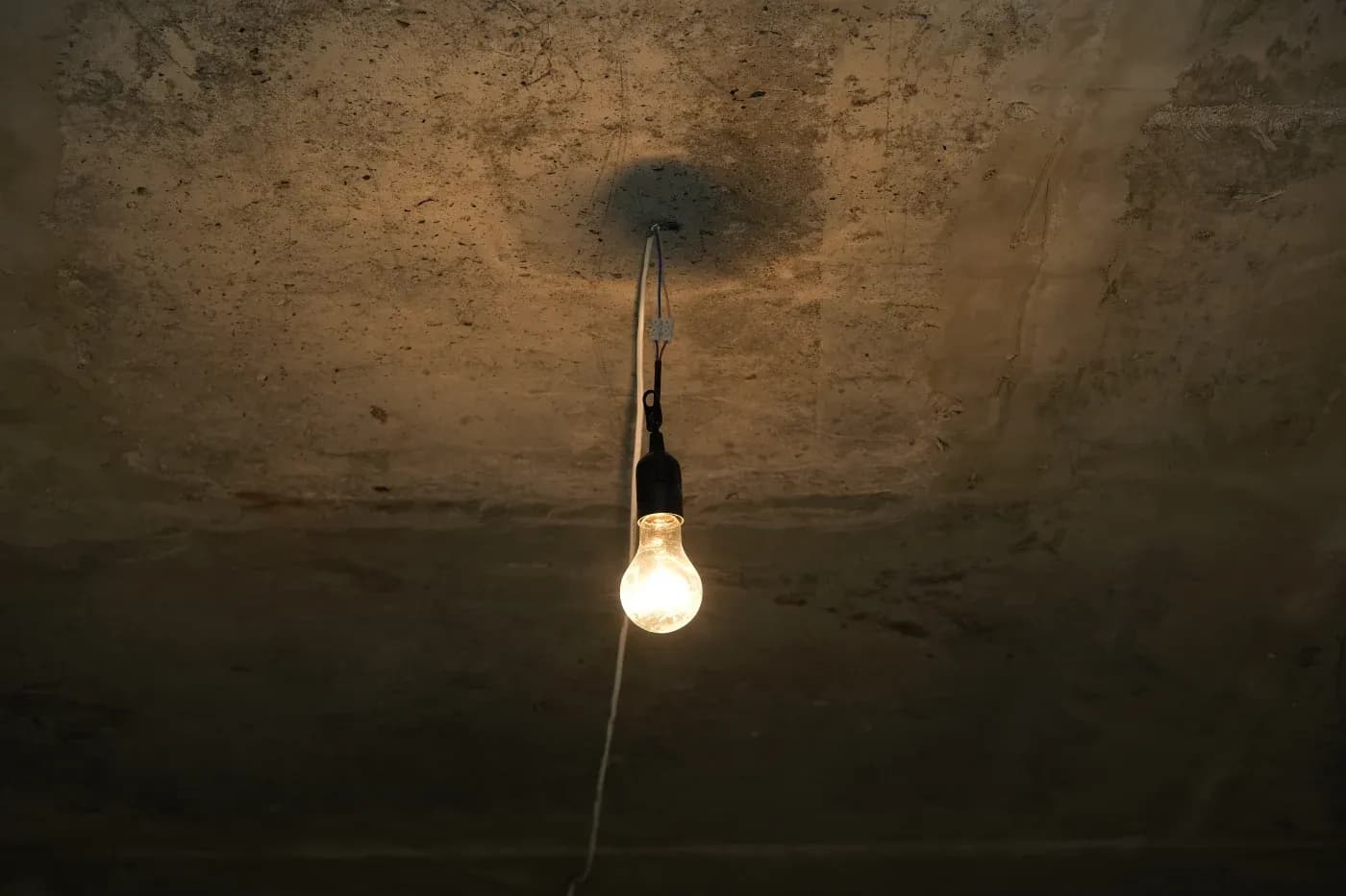 A single exposed light bulb hangs from a weathered, concrete ceiling, casting a dim glow. The setting feels rustic and evokes a sense of simplicity.