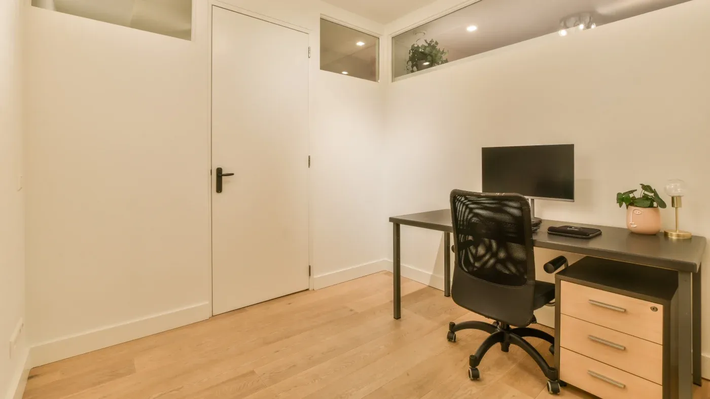Minimalist home office with a black desk, computer monitor, and chair. A small potted plant and lamp sit on the desk. Light wood flooring and white walls create a clean, calm atmosphere.