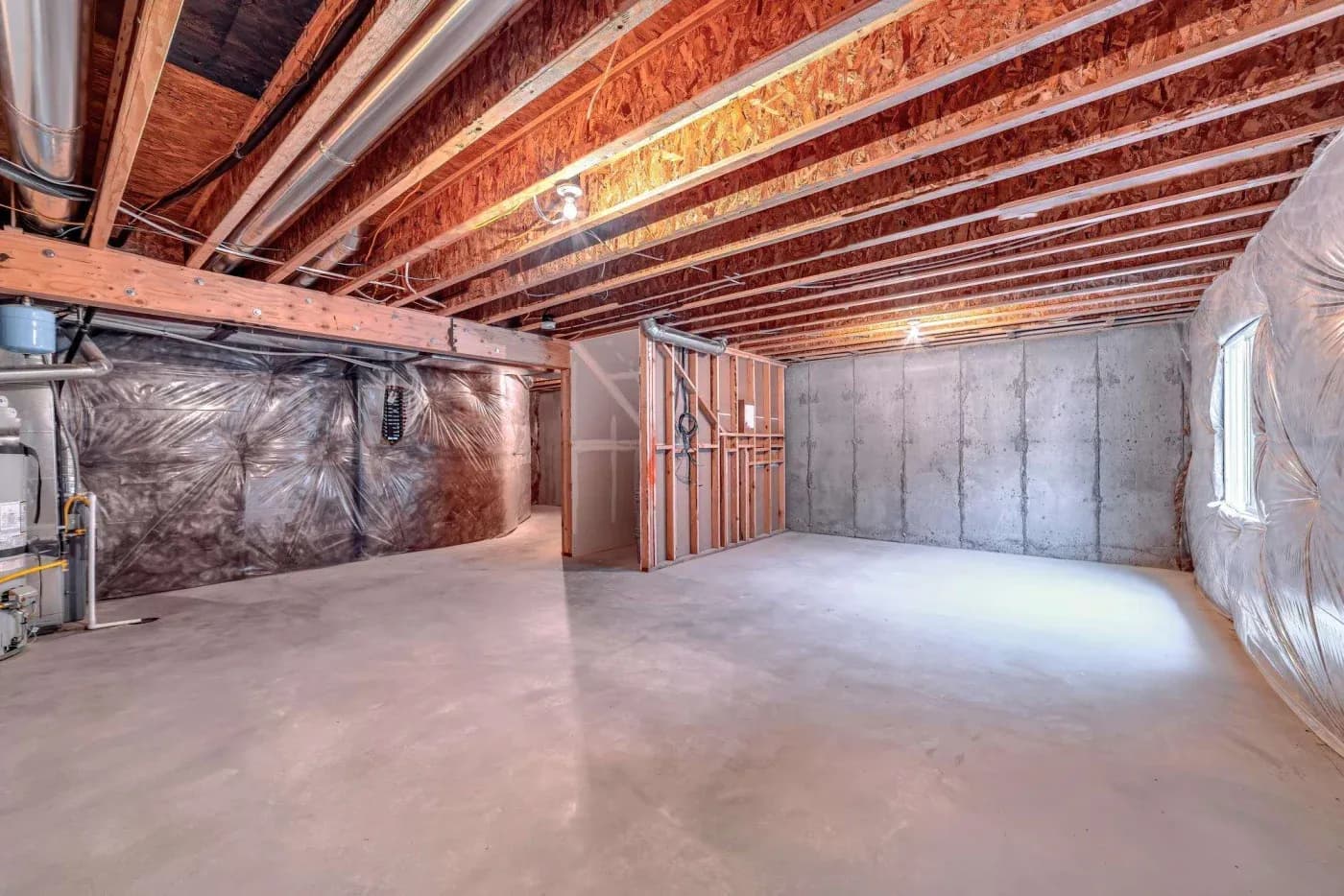 A large, unfinished basement with exposed wooden beams and concrete walls. The space is empty, with plastic-wrapped insulation visible on the sides. Bright overhead lighting illuminates the concrete floor, creating a clean, spacious atmosphere.