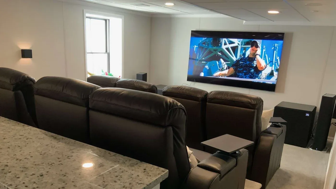 Home theater room with six leather recliners facing a large wall-mounted screen showing an action scene. Soft lighting creates a cozy, cinema-like ambiance.