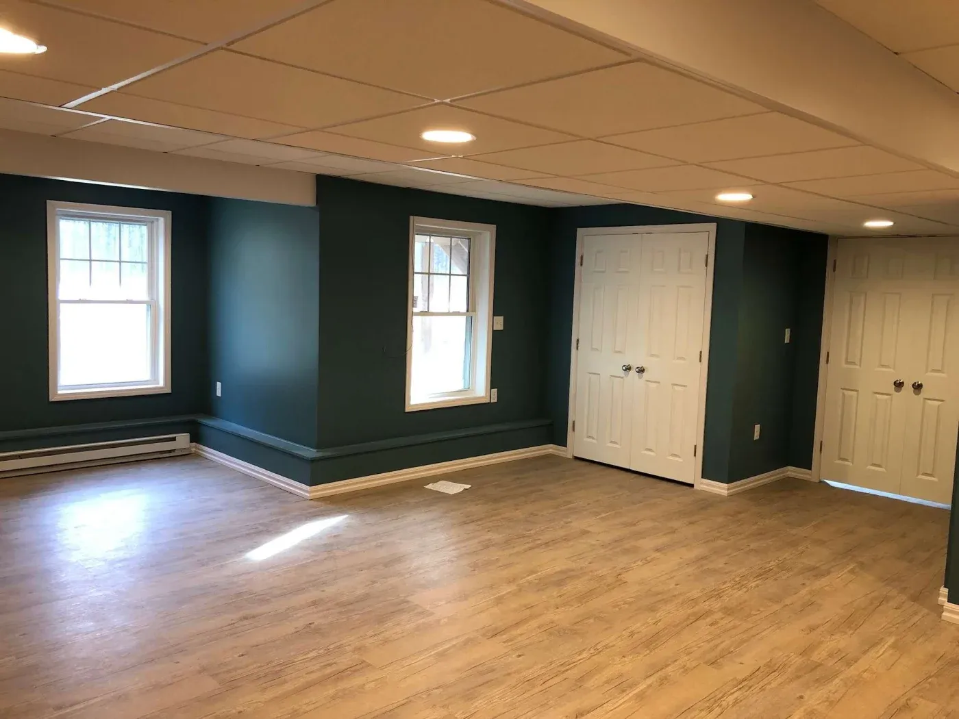 Spacious room with dark teal walls, light wood flooring, and two large windows. White double doors and recessed lighting create a modern, airy feel.