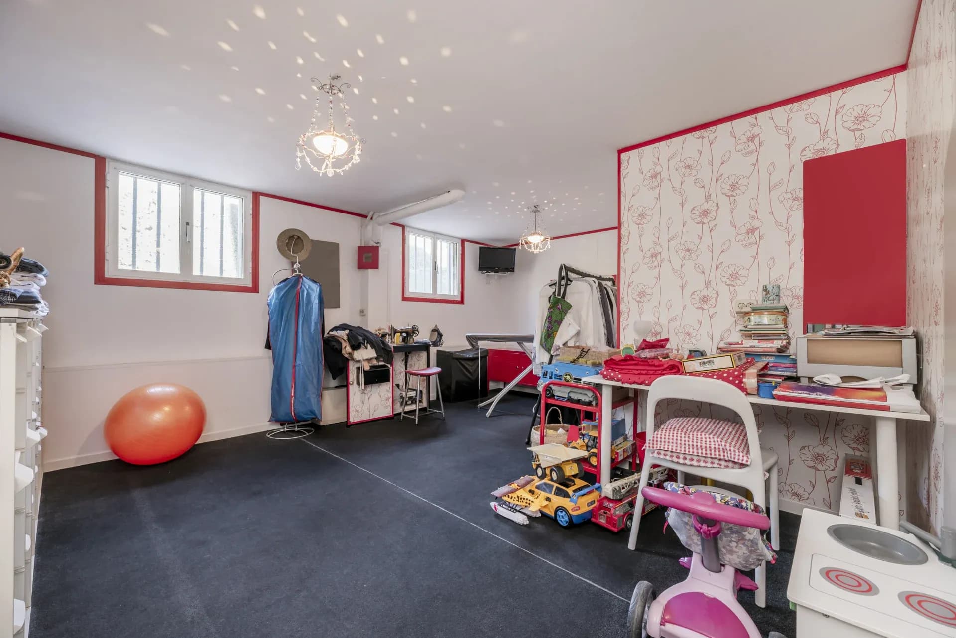 A spacious, colorful room with toys, an orange exercise ball, a small table, and a chandelier. Two windows let in light, creating a playful atmosphere.