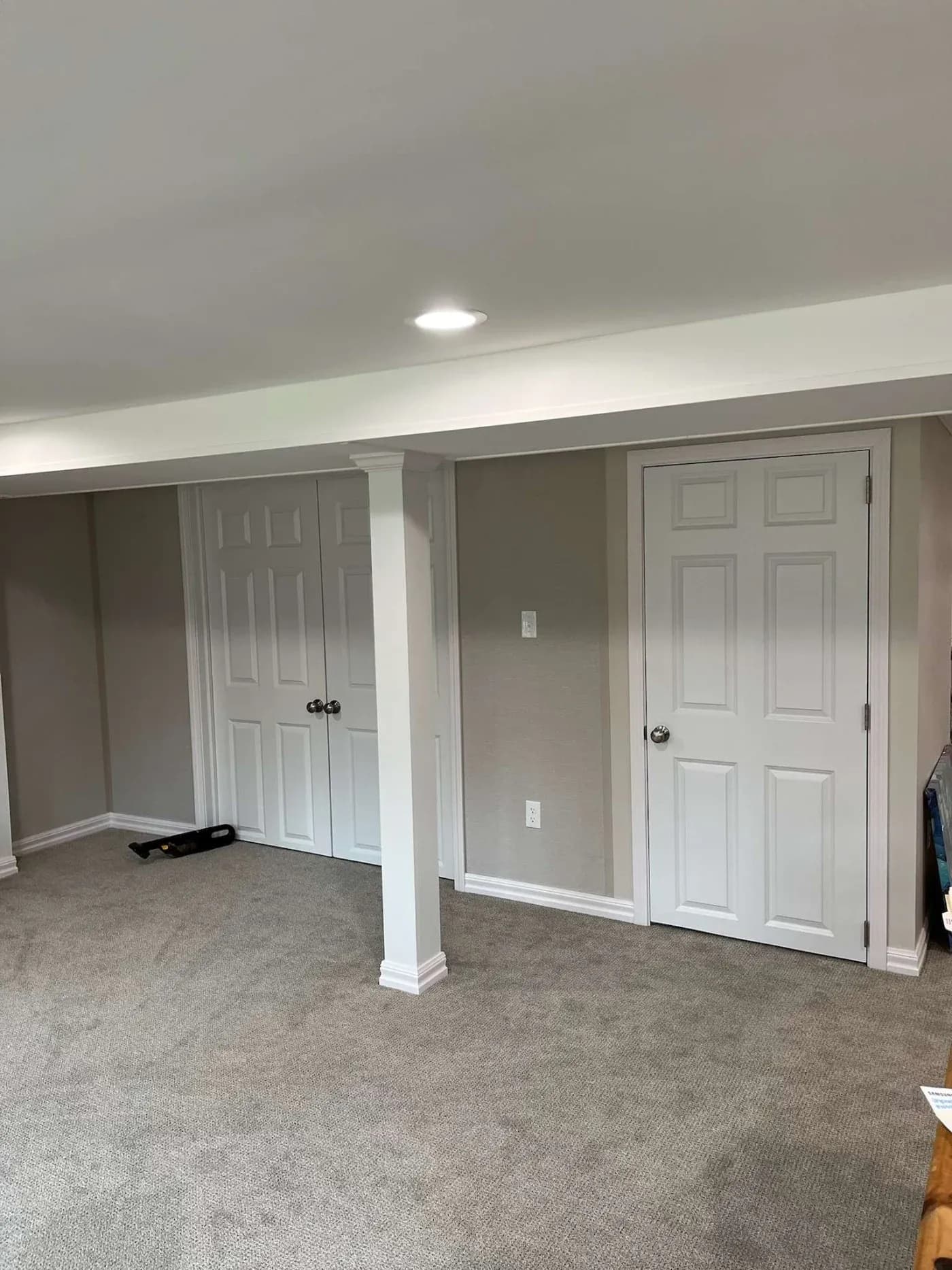 A basement room with light gray carpet, two closed white doors, and a supporting column. The lighting is bright and the atmosphere is clean and minimalistic.