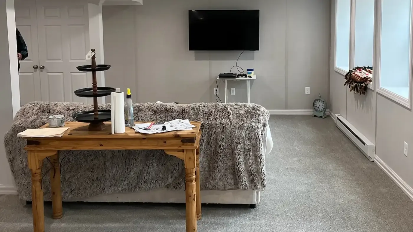 A cozy basement living space with a TV on a gray wall, fuzzy sofa with a wooden console table behind, holding papers and decor. A rug and clock add charm.