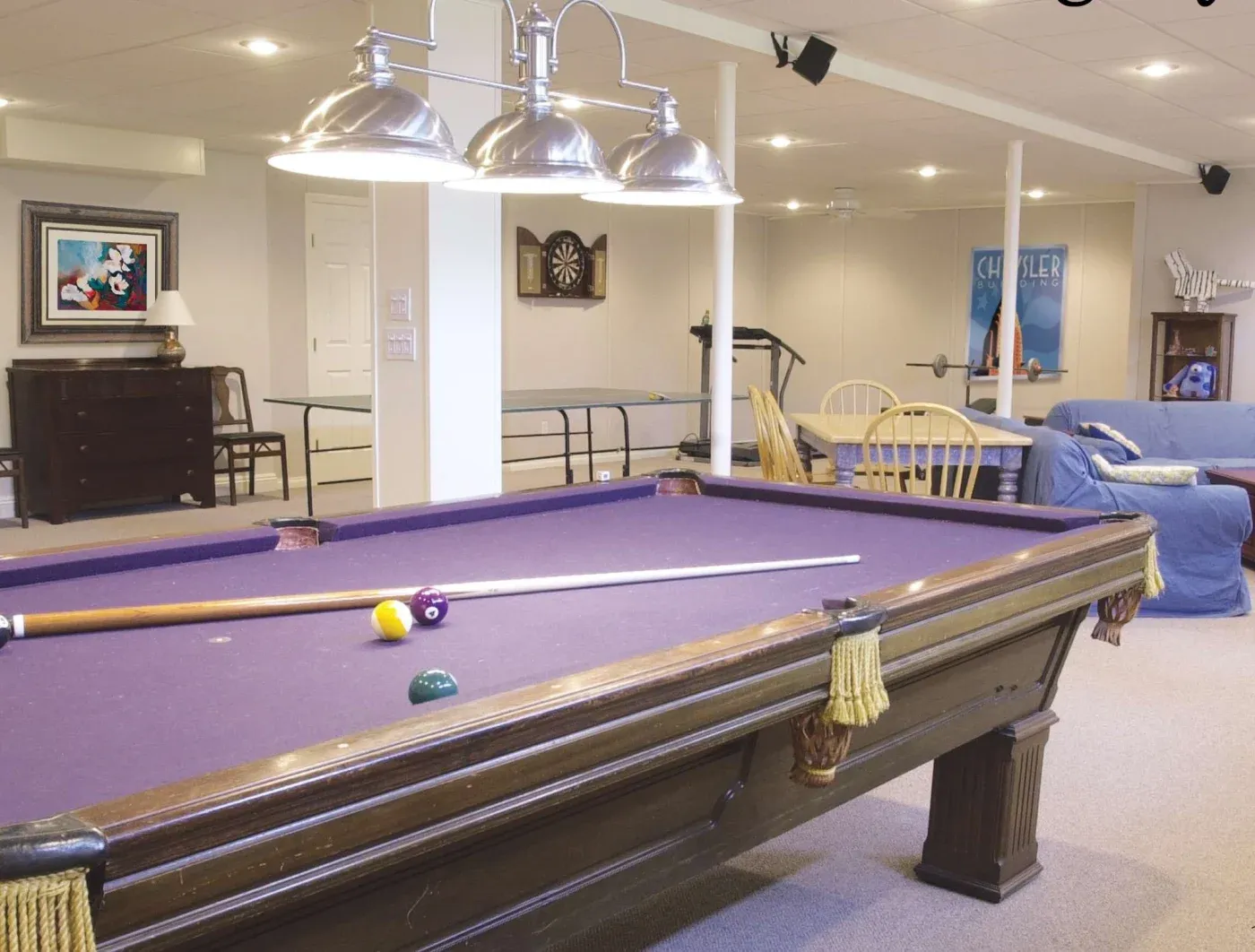 A cozy game room with a purple pool table in the foreground, surrounded by modern light fixtures. In the background, there’s a dartboard, a sofa, chairs, and framed art, creating a relaxed, inviting atmosphere.