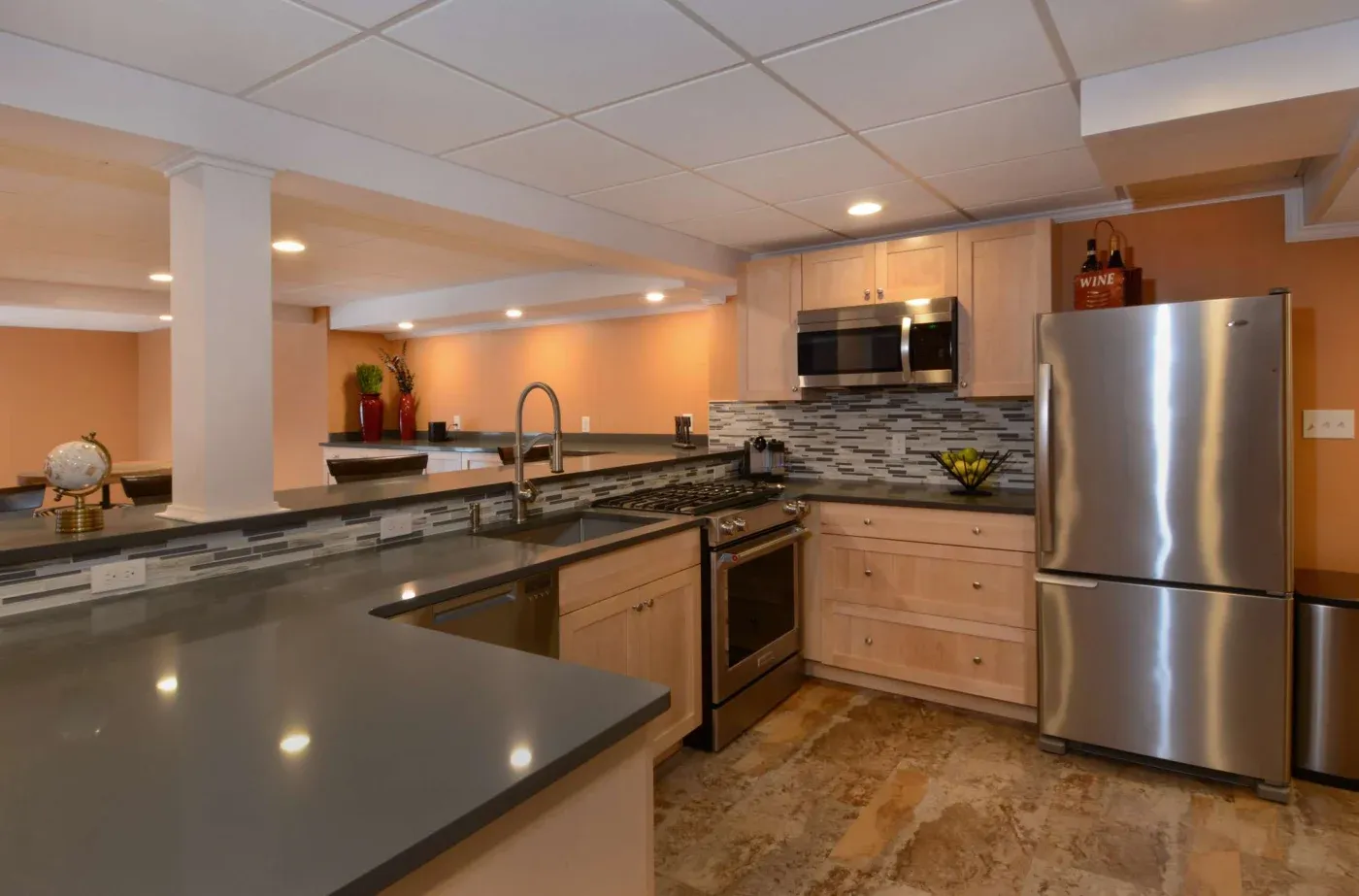 Modern kitchen with stainless steel appliances, light wood cabinets, and dark countertops. Tile flooring, orange walls, and under-cabinet lighting add warmth.