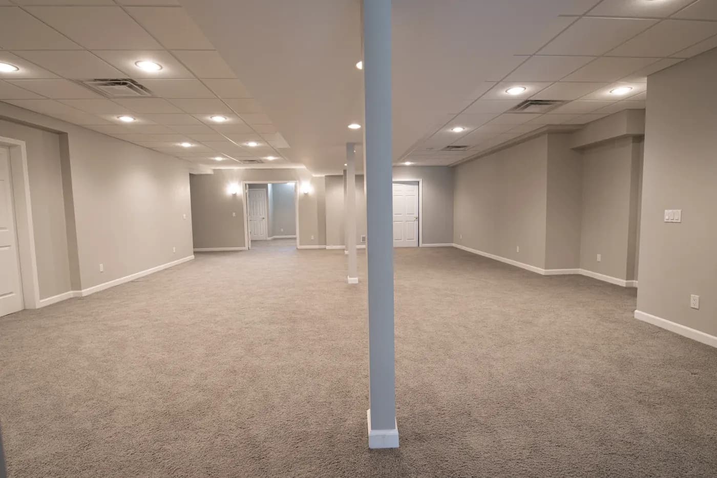 Spacious, brightly lit basement with gray walls, carpeted floor, and white ceiling tiles. Two columns divide the room, creating an open, inviting atmosphere.