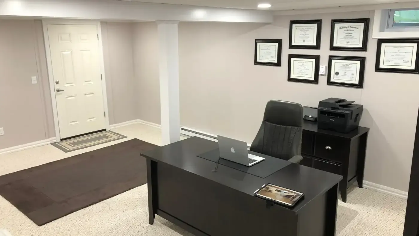 A minimalist office with a dark desk and chair, a laptop, and a printer. Five framed certificates adorn the walls, exuding a professional atmosphere.
