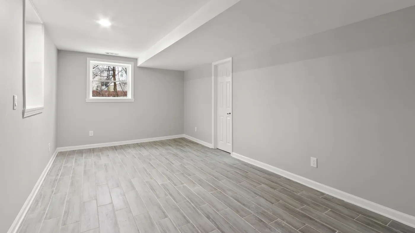A spacious, empty room with light gray walls and wood-look tile flooring. A single window brightens the space, and a closed white door is on the right wall.