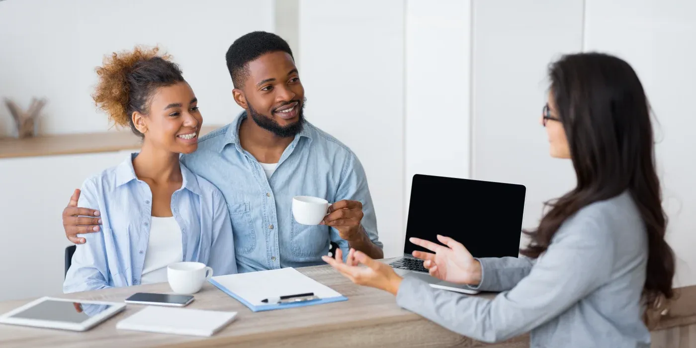 A couple sits at a table, smiling and holding coffee cups, engaged in conversation with a businesswoman opposite them. A laptop and documents are on the table.