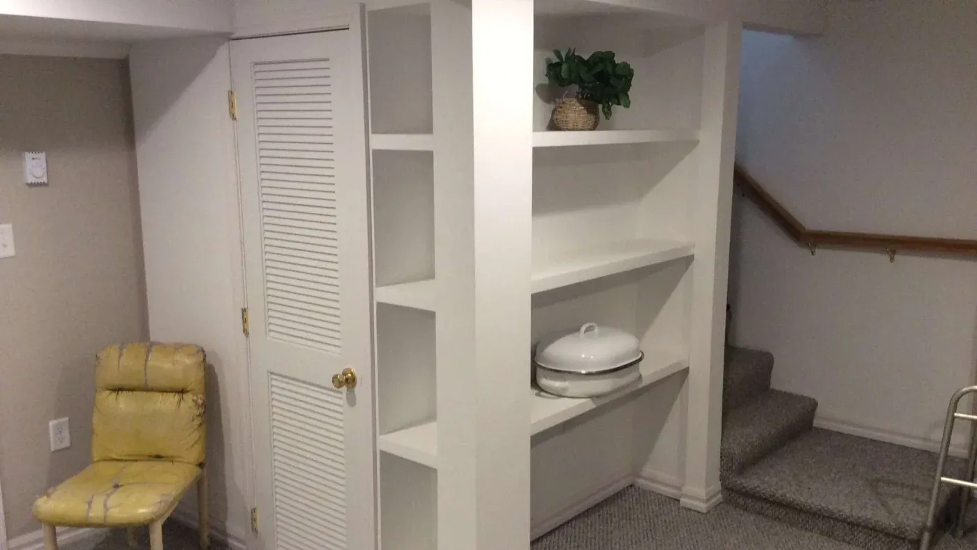 A small basement with white shelving, displaying a potted plant and casserole dish. Beige walls, a worn yellow chair, and carpeted stairs create a cozy atmosphere.