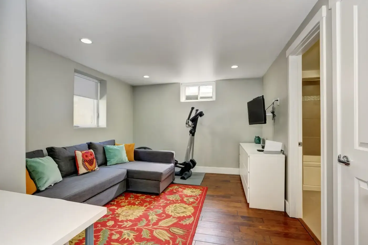 A cozy, modern room with gray walls features a gray sectional sofa adorned with colorful pillows, a vibrant red floral rug, a stationary bike, and a wall-mounted TV. The room has recessed lighting, wooden floors, and a white cabinet beside a bathroom door, creating a comfortable and inviting atmosphere.