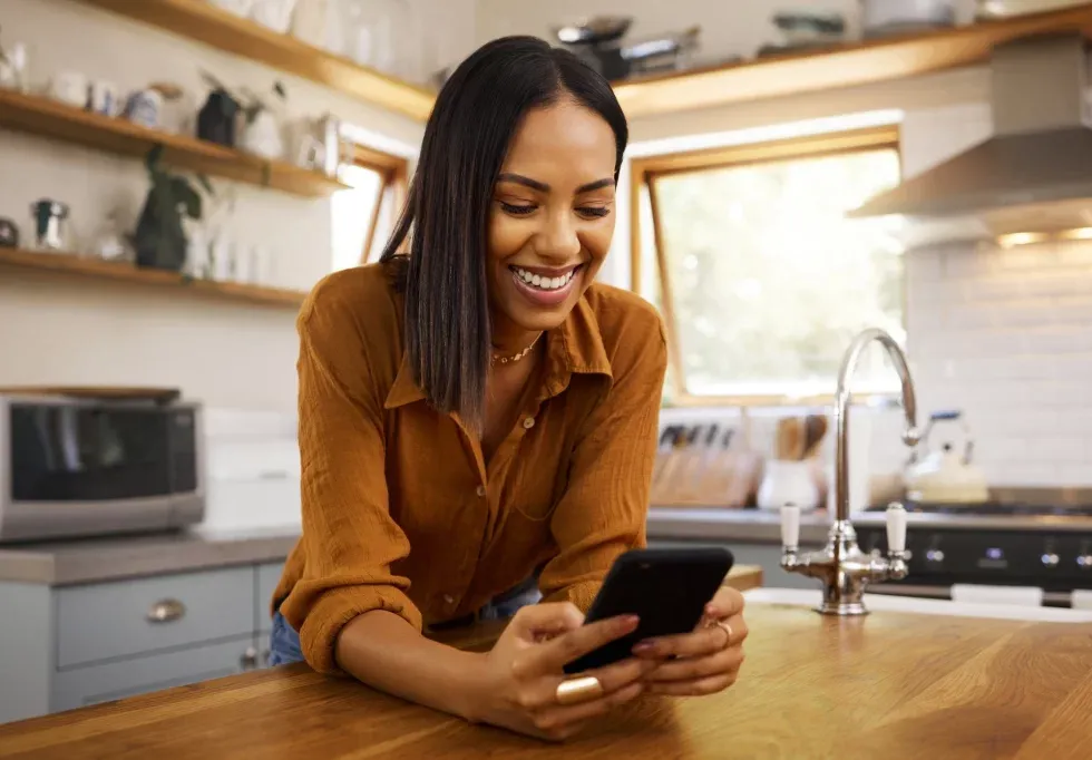 A woman in a cozy kitchen smiles while looking at her phone. She leans on a wooden countertop, creating a warm and relaxed atmosphere.