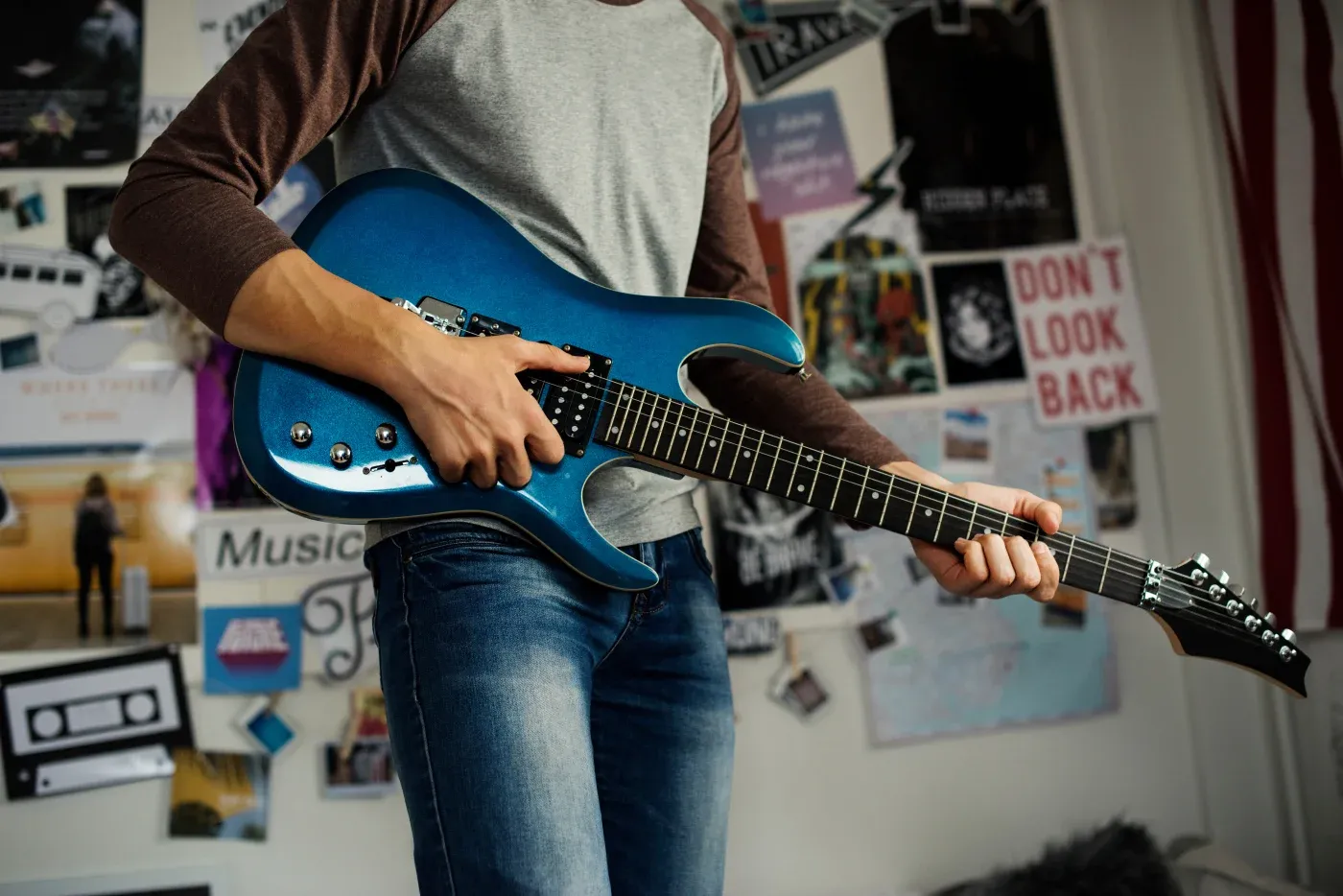 Person playing a blue electric guitar in a room with collage-covered walls. The atmosphere is creative and musical, with a casual vibe.