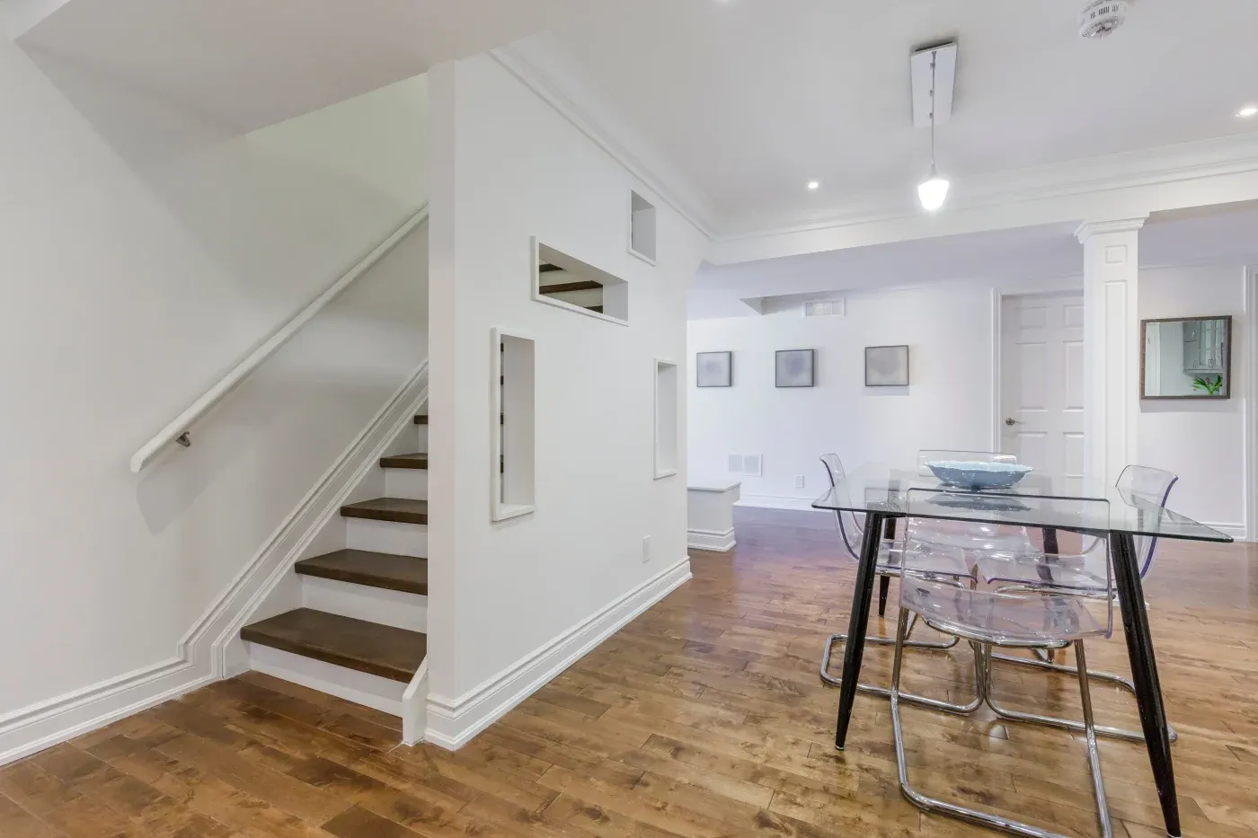 Modern dining area with glass table and clear chairs on wooden floor. Nearby, a staircase with white walls and framed pictures creates a minimalist feel.