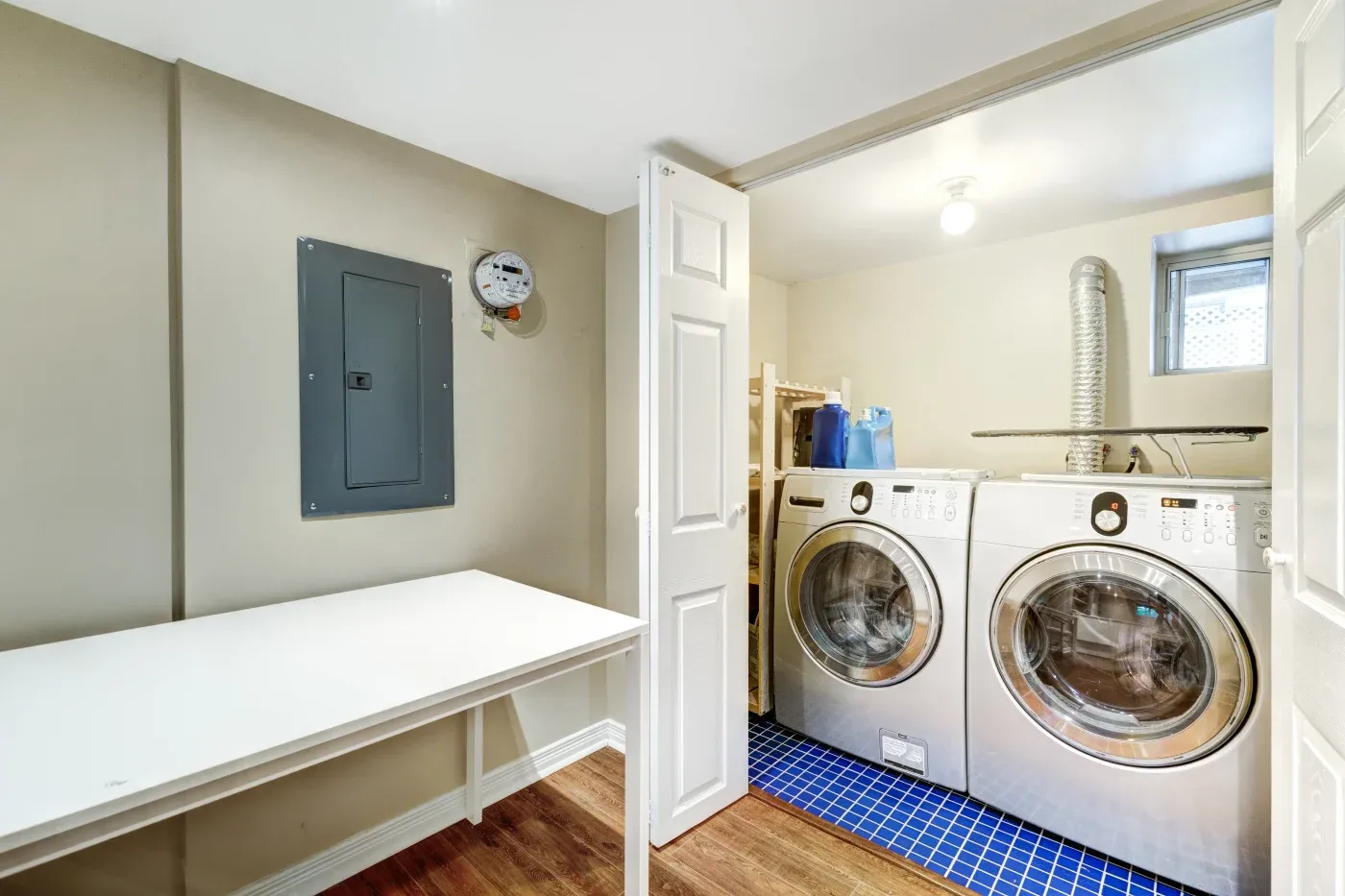 A modern laundry room featuring a washing machine and dryer on blue tile flooring. A folding table is adjacent, with beige walls and wood flooring.