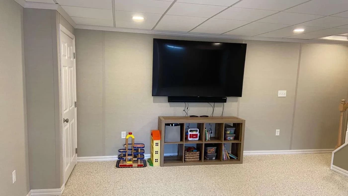 A basement room with beige walls and carpet features a large TV mounted above a wooden shelf. The shelf holds toys and books, creating a cozy, family-friendly space.