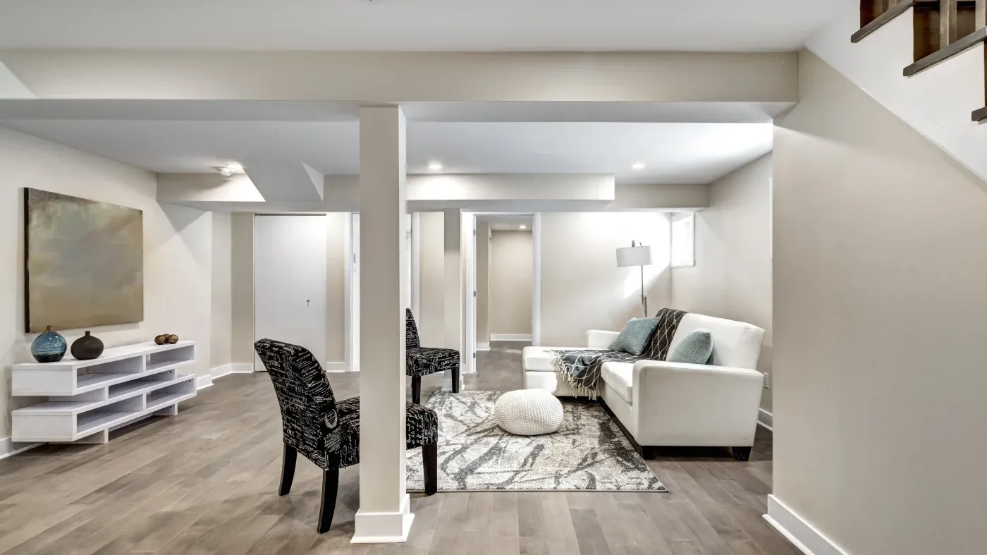 Modern basement living area with neutral tones, featuring a white sofa, patterned chairs, a rug, and minimalist decor. Soft lighting creates a cozy ambiance.